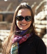 Foto da professora Alessandreia sorrindo. Ela tem cabelos longos na cor escura, pele branca. Usa óculos de armação preta e um cachecol nos tons rosa, roxo e preto. O fundo da imagem é borrado em tons de marrom e bege.