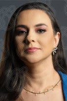 Foto da professora Kamila. Ela tem cabelos longos na cor escura, pele branca. Usa maquiagem em tons de rosa, um brinco de folhas, um colar dourado e uma blusa azul. O fundo da imagem é cinza.
